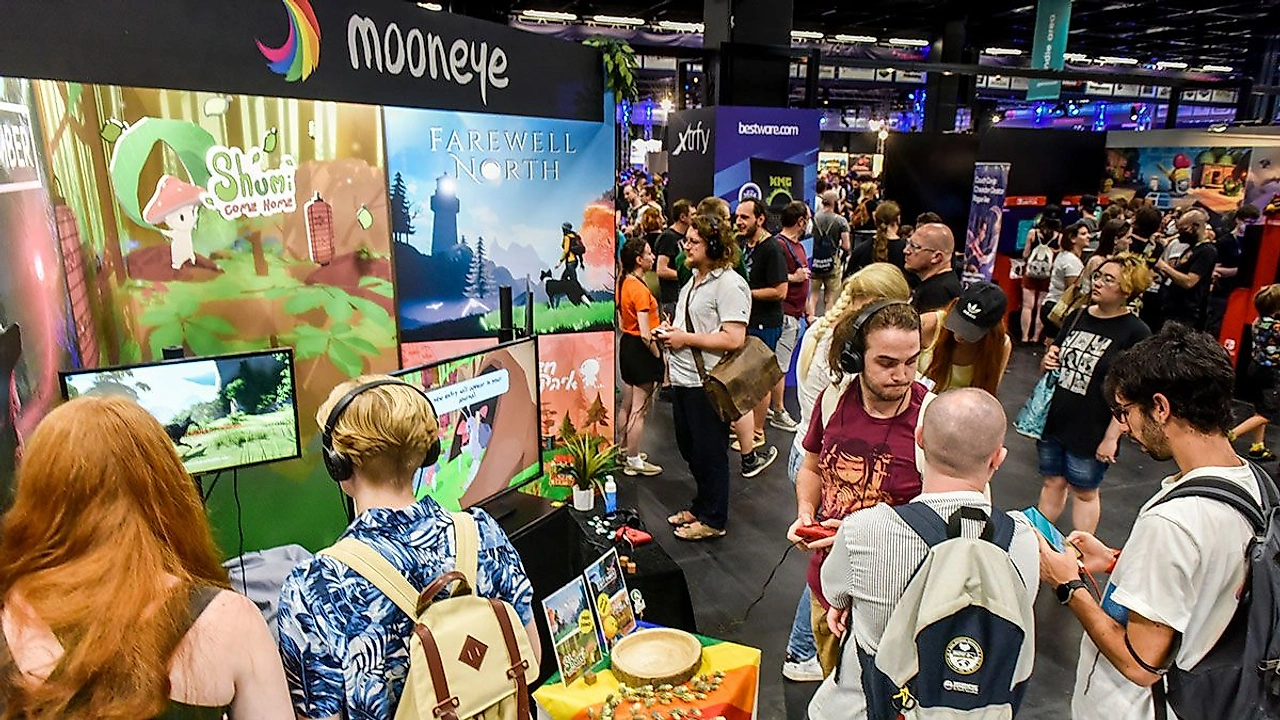 Kalabalık bir fuar alanında Mooneye standı önünde oyun oynayan ve sohbet eden katılımcılar; arka planda "Shumi Come Home" ve "Farewell North" oyun afişleri, renkli dekorlar ve farklı stantlar dikkat çekiyor.